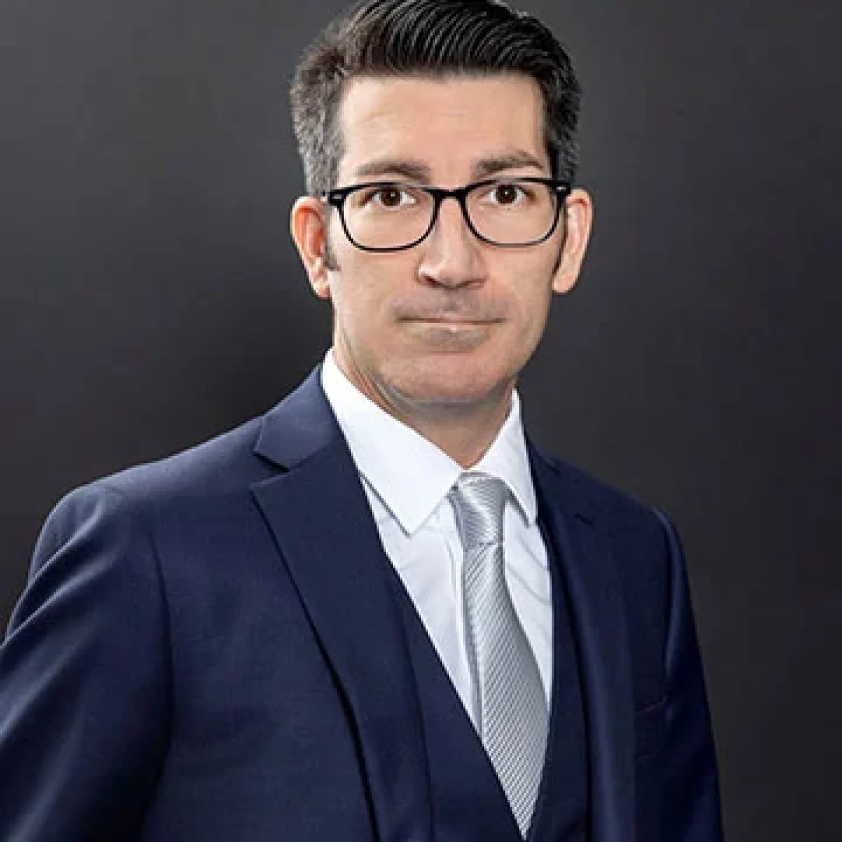 Man wearing glasses, a navy blue suit, white dress shirt, and light gray tie, posing against a plain dark background.