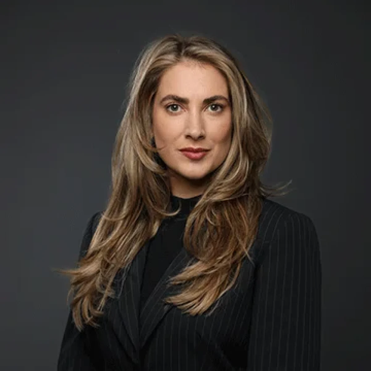 A woman with long, wavy blonde hair wearing a black pinstripe blazer poses in front of a plain dark background.