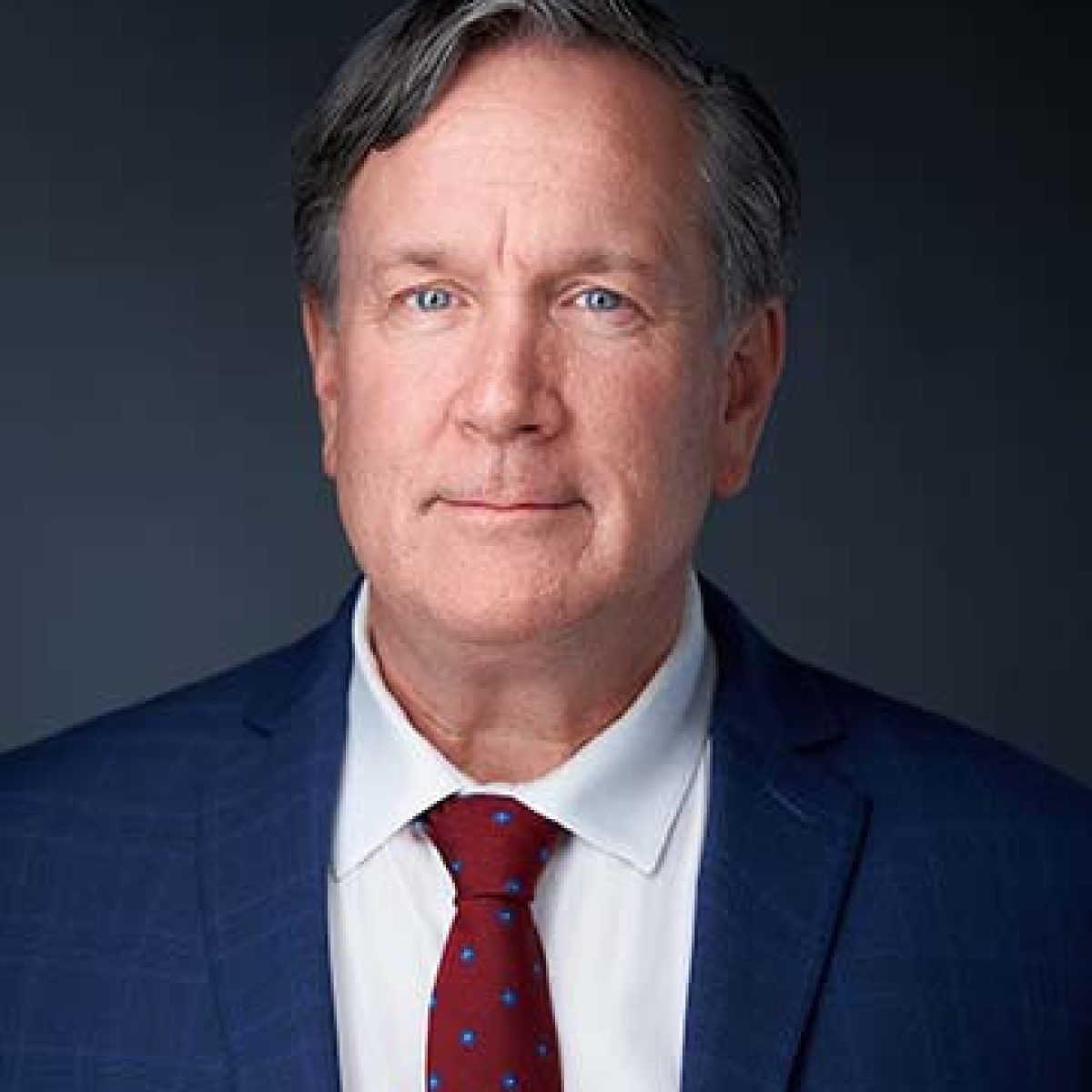 Middle-aged man with short gray hair wearing a navy suit, white shirt, and red tie, posing against a dark background.